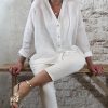 Nancy zittend op een bank draagt witte linnen blouse met V-hals en opgerolde mouwen, gecombineerd met ecru jeans en gouden sandalen.